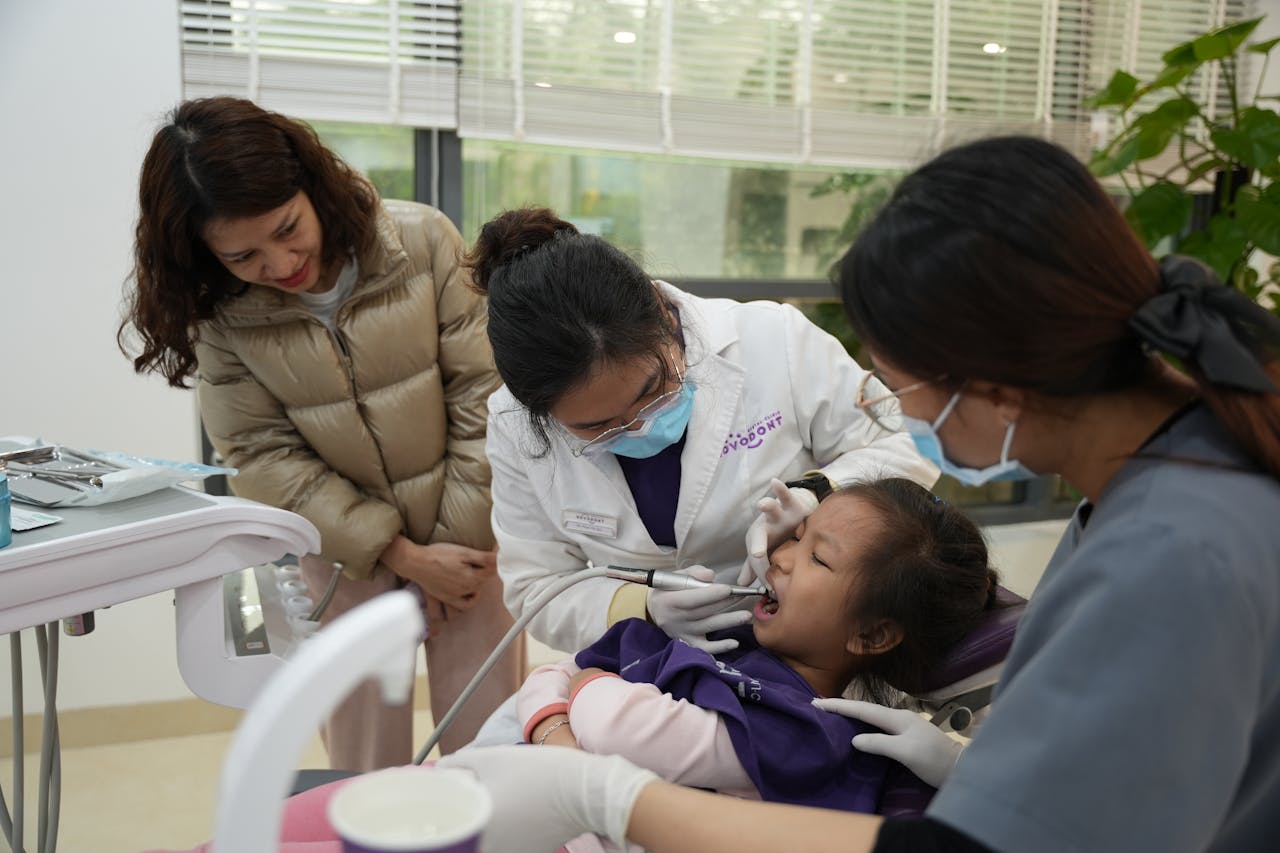 A dentist examines a young girls teeth, with a parent and assistant observing in a modern clinic.